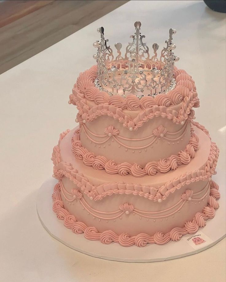 a three tiered cake with pink frosting and a crown on top is sitting on a table