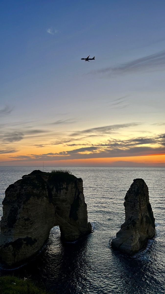 an airplane is flying over the ocean with two rocks in the foreground and another rock outcropping on the other side