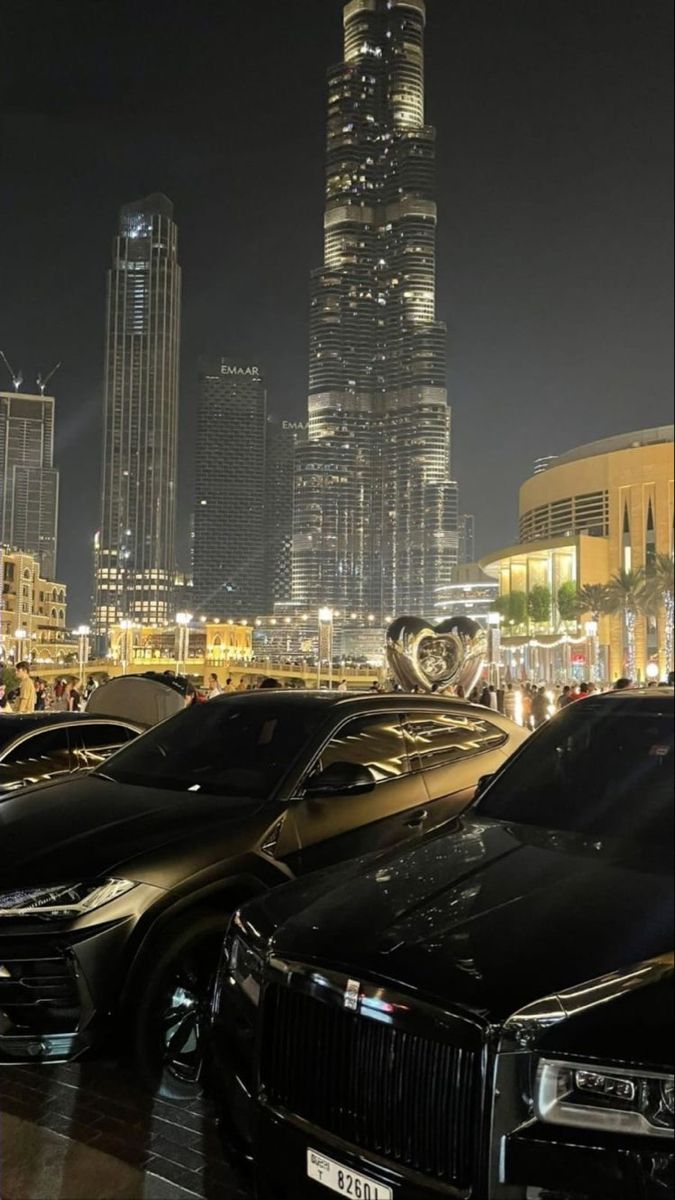 several cars parked in front of a tall building at night with lights shining on the buildings