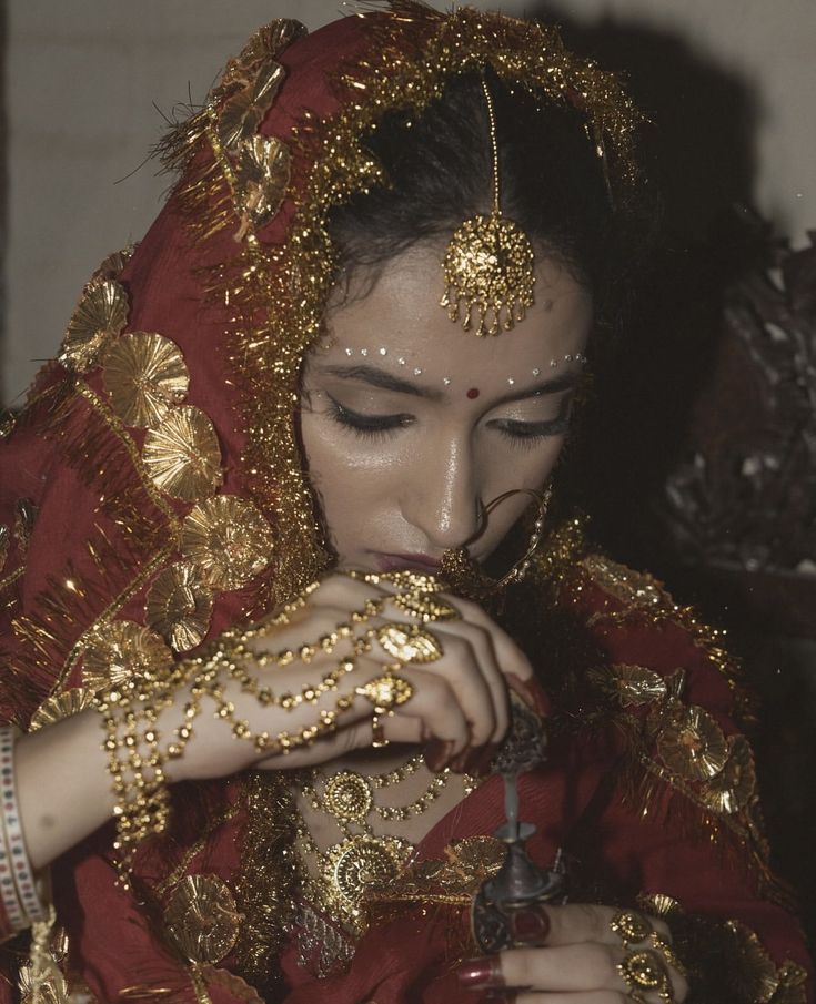 a woman dressed in red and gold with jewelry on her head, holding an object to her mouth