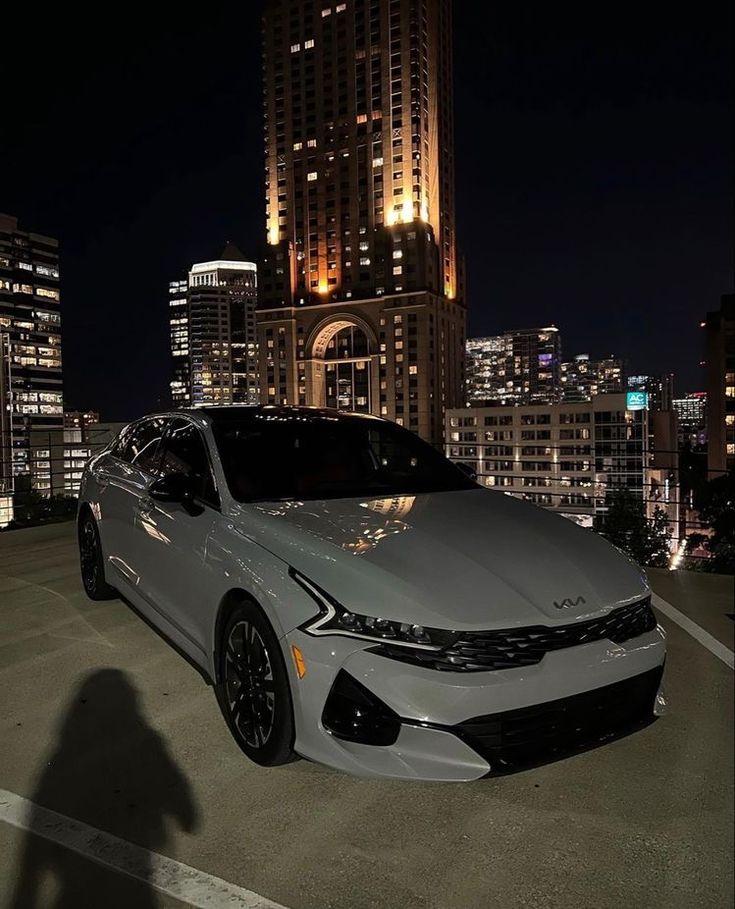 a white car parked in front of a city at night with lights shining on the buildings