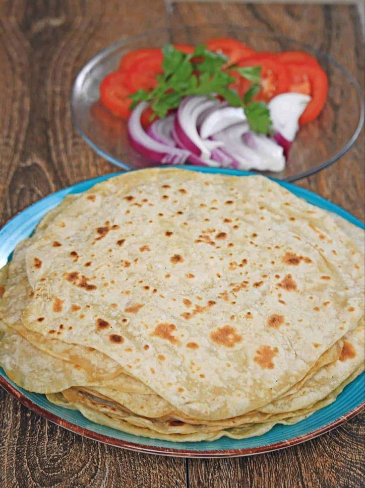 three flat breads on a blue plate next to a bowl of tomatoes and onions