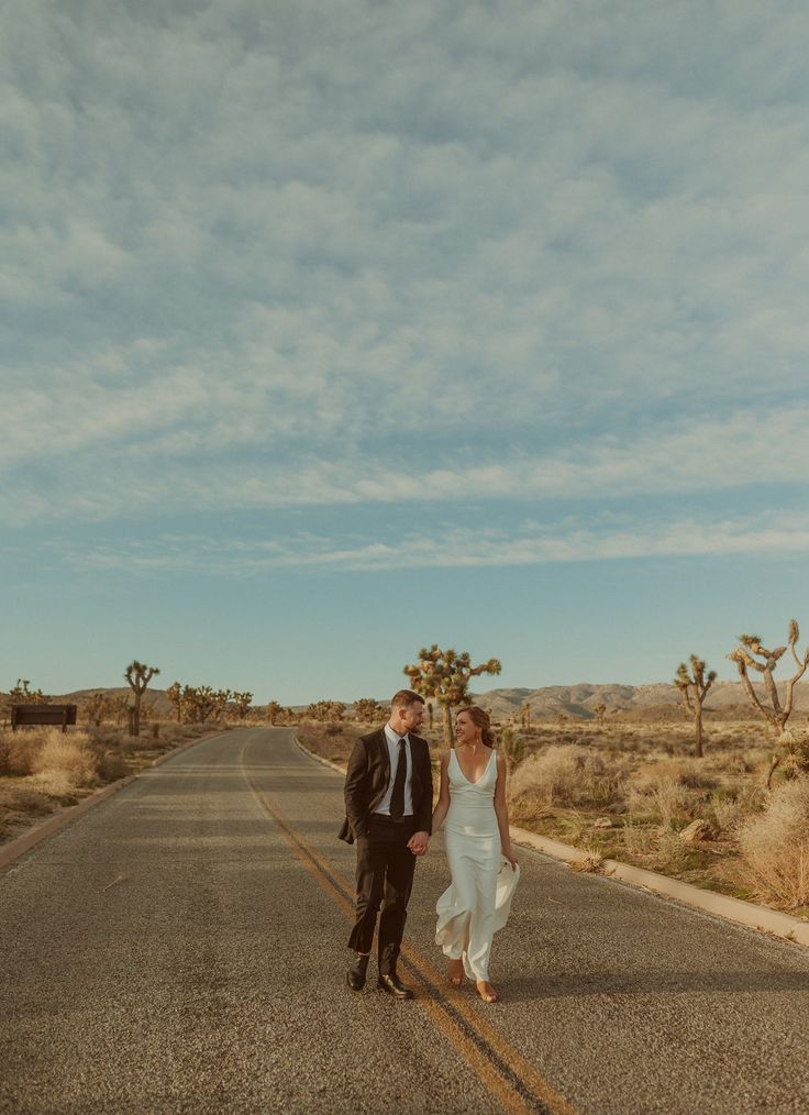 a bride and groom walking down the middle of an empty road with cacti in the background