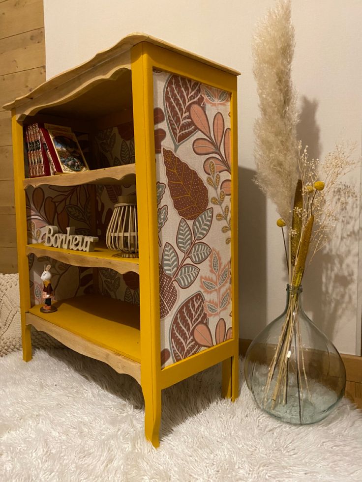 a yellow book shelf sitting on top of a white fluffy rug next to a vase