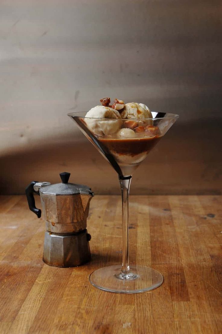 a martini glass filled with ice cream and toppings next to a coffee pot on a wooden table