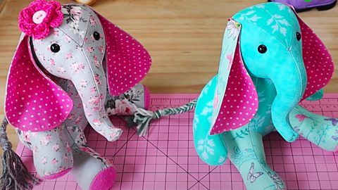 two stuffed animals sitting on top of a pink cutting board next to scissors and sewing needles