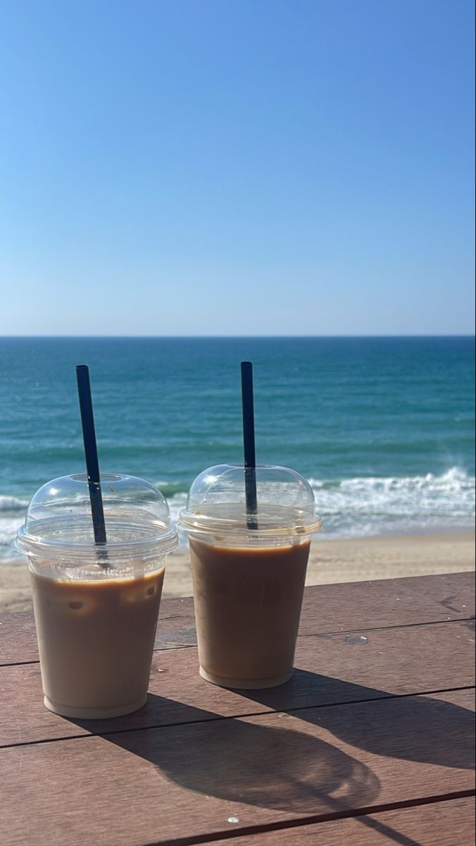 two drinks sitting on top of a wooden table next to the ocean