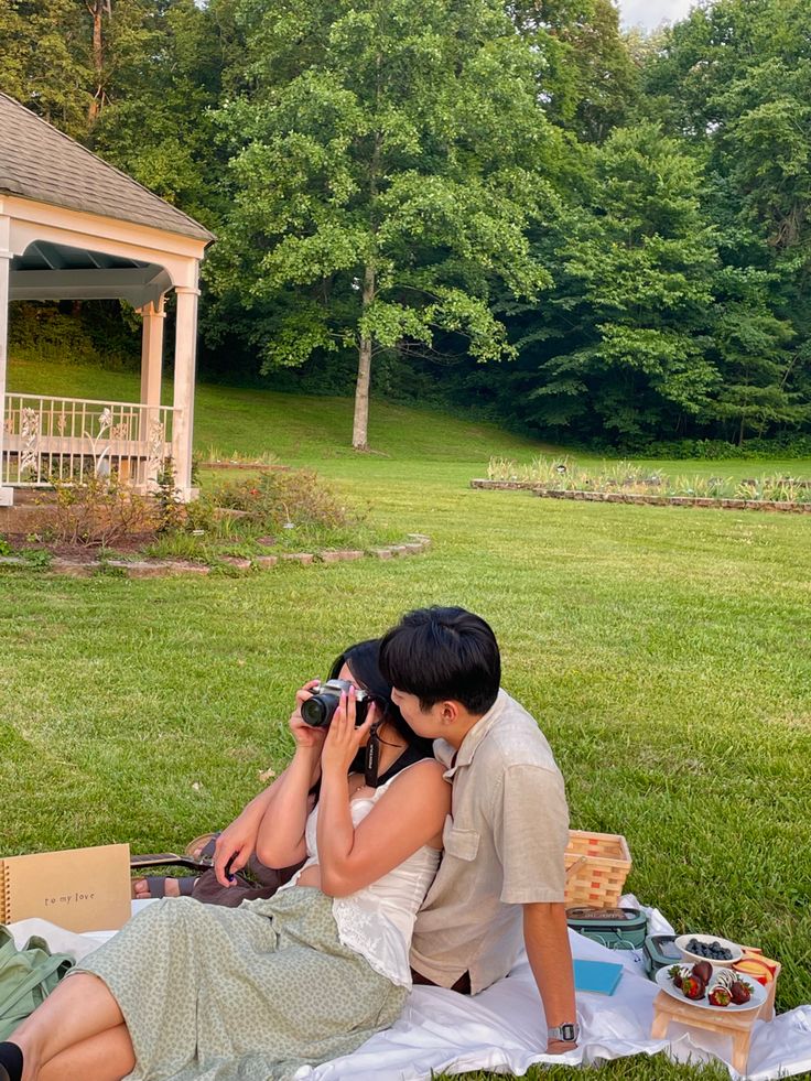 a man and woman sitting on a blanket in front of a gazebo with food