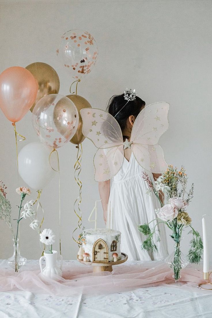 a woman in white dress standing next to balloons and cake with fairy wings on it