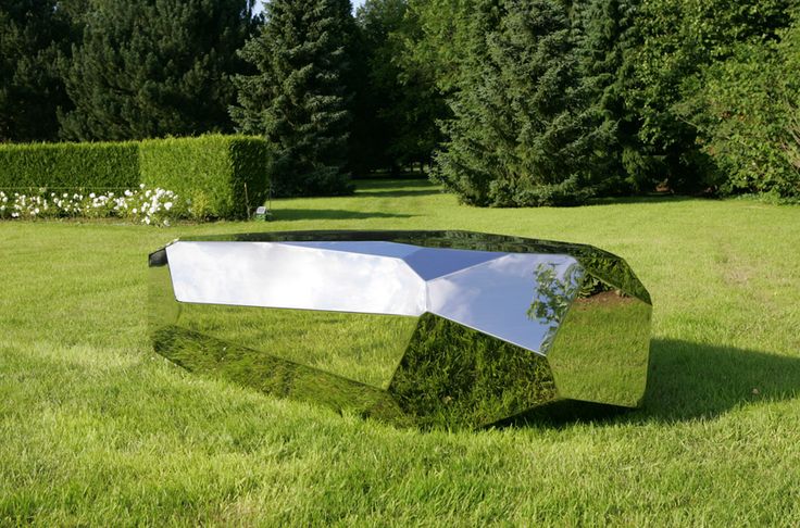 a mirror sculpture sitting on top of a lush green field next to bushes and trees