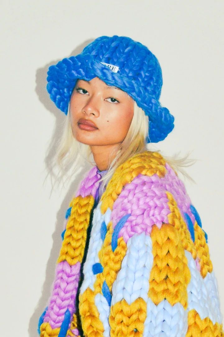 a woman with blonde hair wearing a blue knitted hat and colorful sweater, standing in front of a white wall