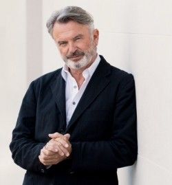 a man standing next to a white wall with his hands folded in front of him