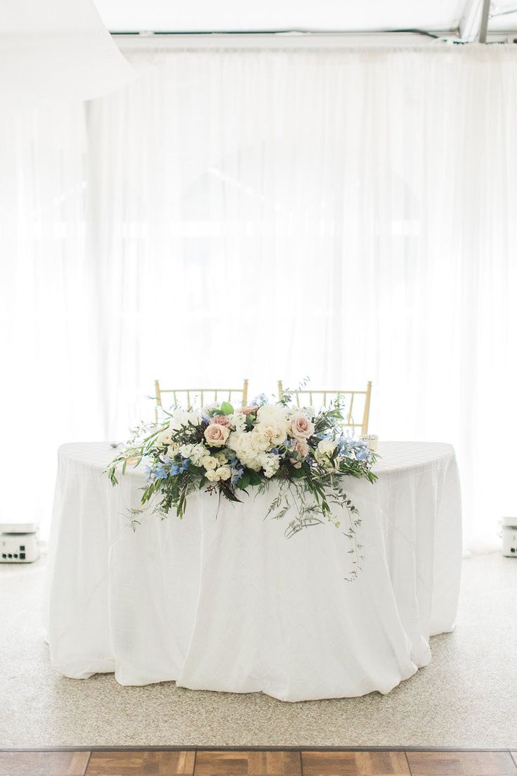 a table with flowers and greenery on it in front of a white drapes