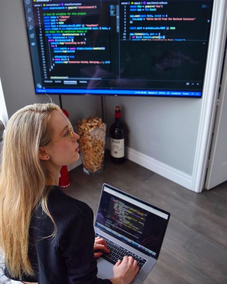 a woman sitting in front of a laptop computer on top of a wooden floor next to a large screen