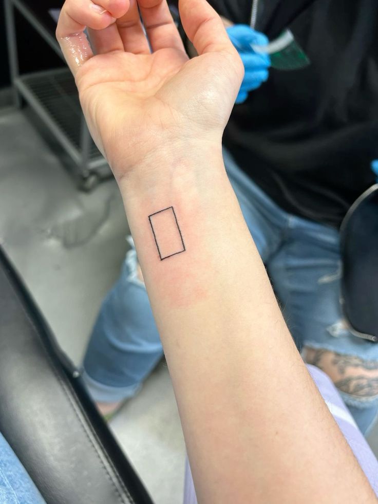 a person with a small square tattoo on their left wrist and hand in the background