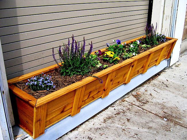 a wooden planter filled with lots of flowers next to a house wall on the sidewalk