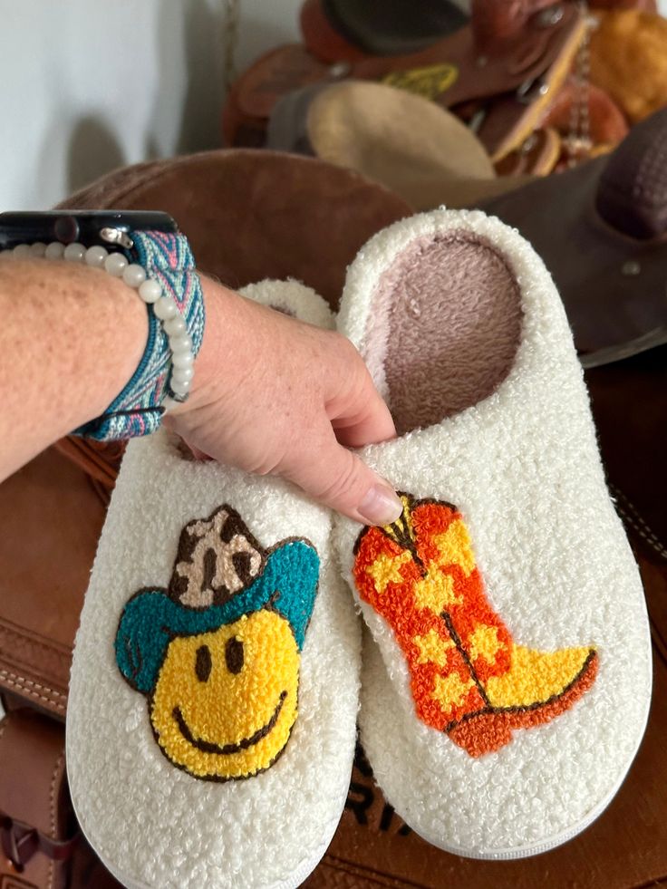 Here’s some fun slippers for the Cowgirls and Cowgirls at heart! These comfy slippers feature a Smiley Face Cowboy and Cowboy Boots. They're incredibly comfy, extra cozy, and feature a rubber bottom ideal for indoor and outdoor wear.Very limited quantity - don't miss out on these fun slippers! Sizing Information:This item is based on women's sizing, and men should size up for the best fit. Size Chart: 6/7: Length 9" 7/8: Length 9 3/4" 8/9: Length 10" 9/10: Length 10 1/2" 11/12: Length 10 3/4" Be Super Soft Comfy Slippers With Round Toe, Comfy Super Soft Round Toe Slippers, Cute Super Soft Indoor Slippers, Fun Slippers With Soft Sole And Round Toe, Cute Round Toe Slippers For Leisure, Cute Round Toe Slippers For Loungewear, Fun Multicolor Round Toe Slippers, Fun Round Toe Winter Slippers, Fun Round Toe Slippers For Winter