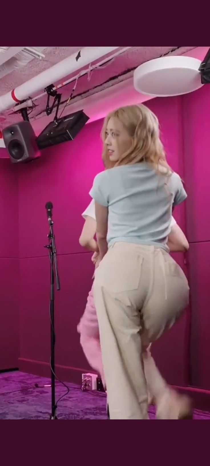 a woman standing in front of a microphone on top of a pink wall next to a speaker