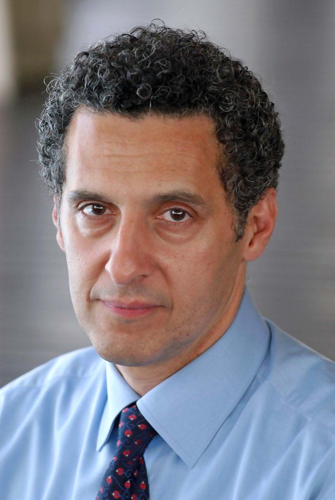 a man with curly hair wearing a blue dress shirt and tie, looking at the camera