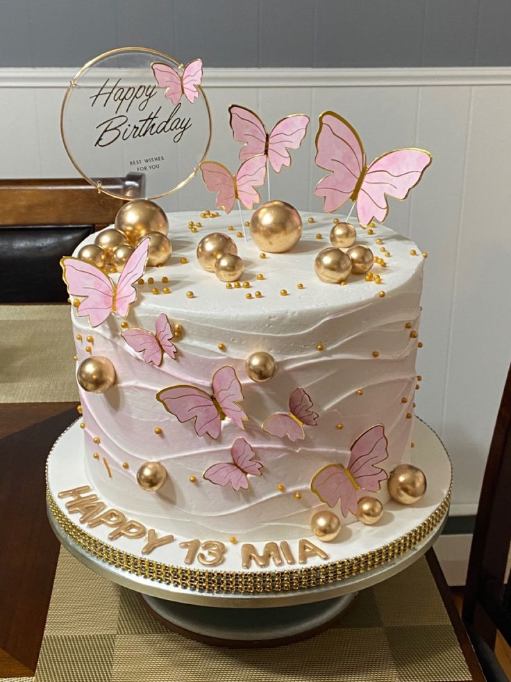 a birthday cake decorated with pink butterflies and gold decorations on a table in front of a happy birthday sign