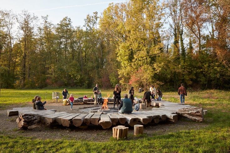 a group of people sitting around a wooden table in the middle of a park area