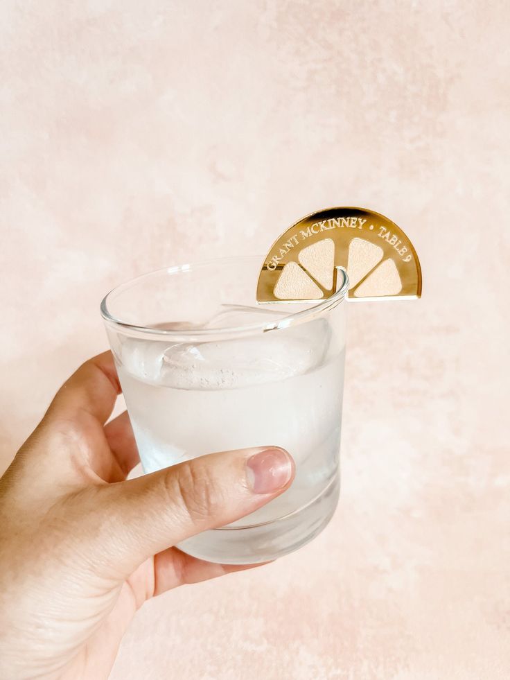 a hand holding a glass filled with water and a lemon slice on top of it