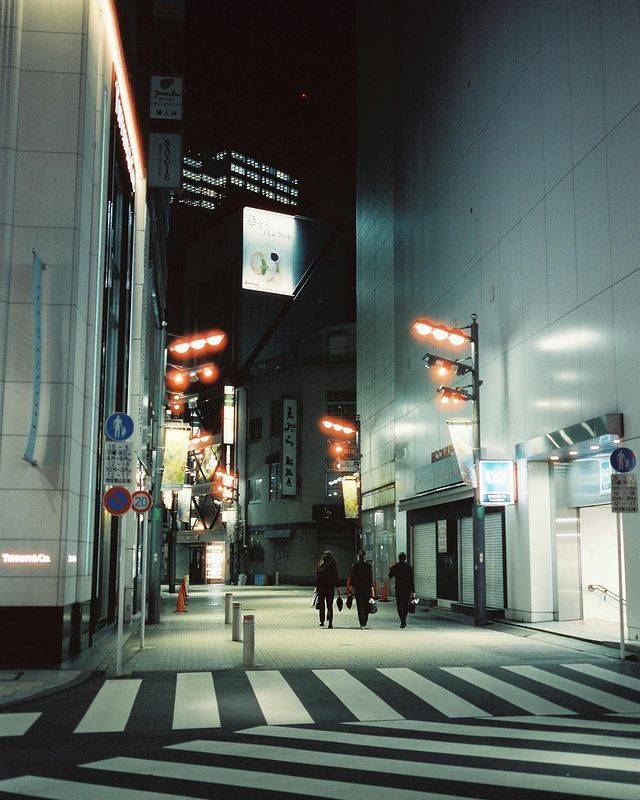 two people walking down the street at night in an urban area with tall buildings and lights