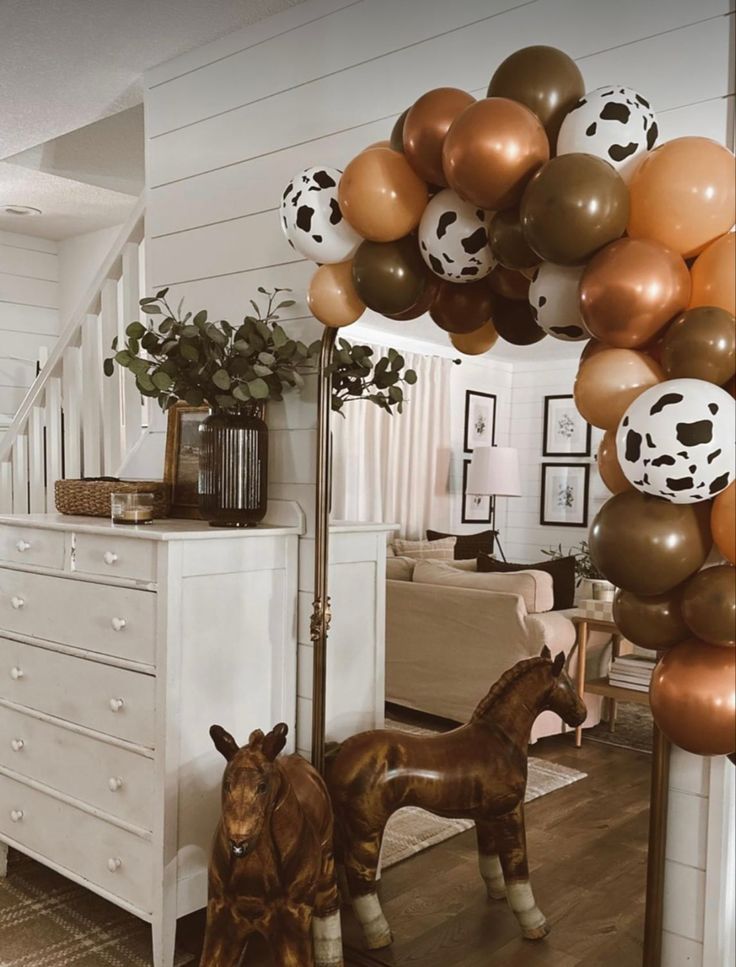 two horses are standing in the living room with balloons on the wall and a cow statue next to them