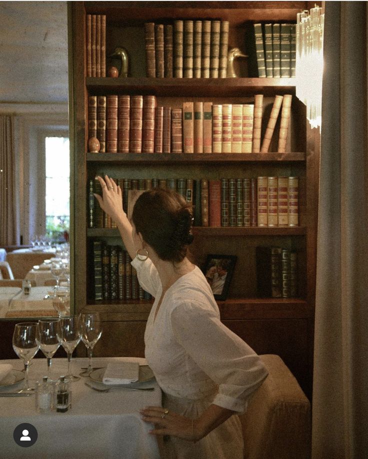 a woman sitting at a table in front of a bookshelf with wine glasses on it