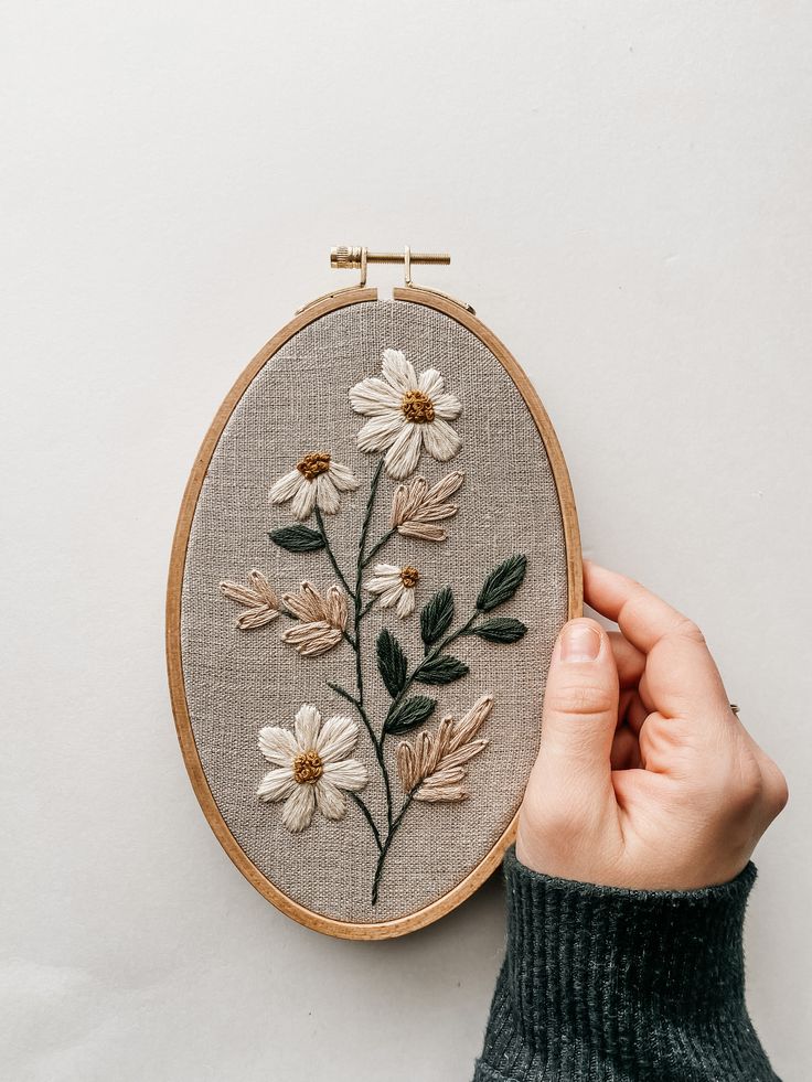 a person holding up a hand embroidered flower on a wall mounted embroidery hoop with white daisies and green leaves