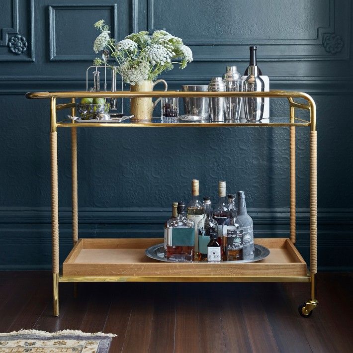 a gold bar cart with bottles and glasses on it in front of a blue wall