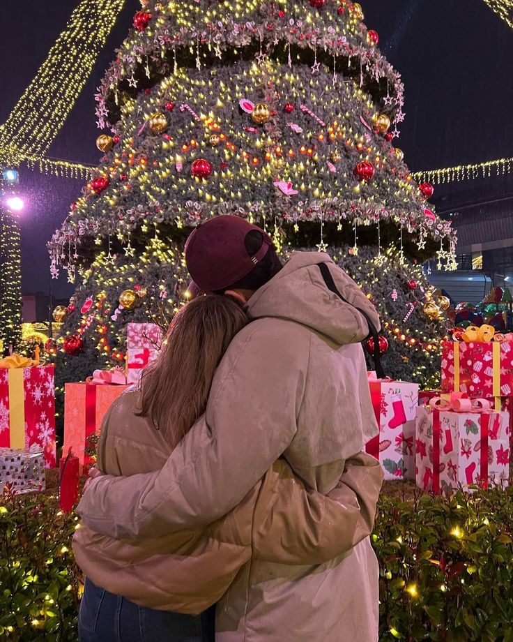 two people standing in front of a christmas tree with presents on it and lights all around