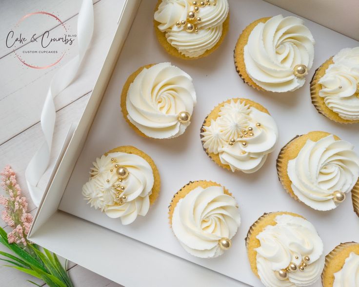 twelve cupcakes with white frosting in a box on a table next to flowers