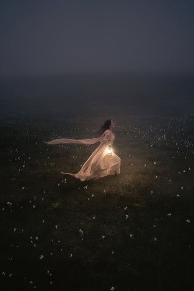 a woman in a white dress is walking through the grass at night with her light on