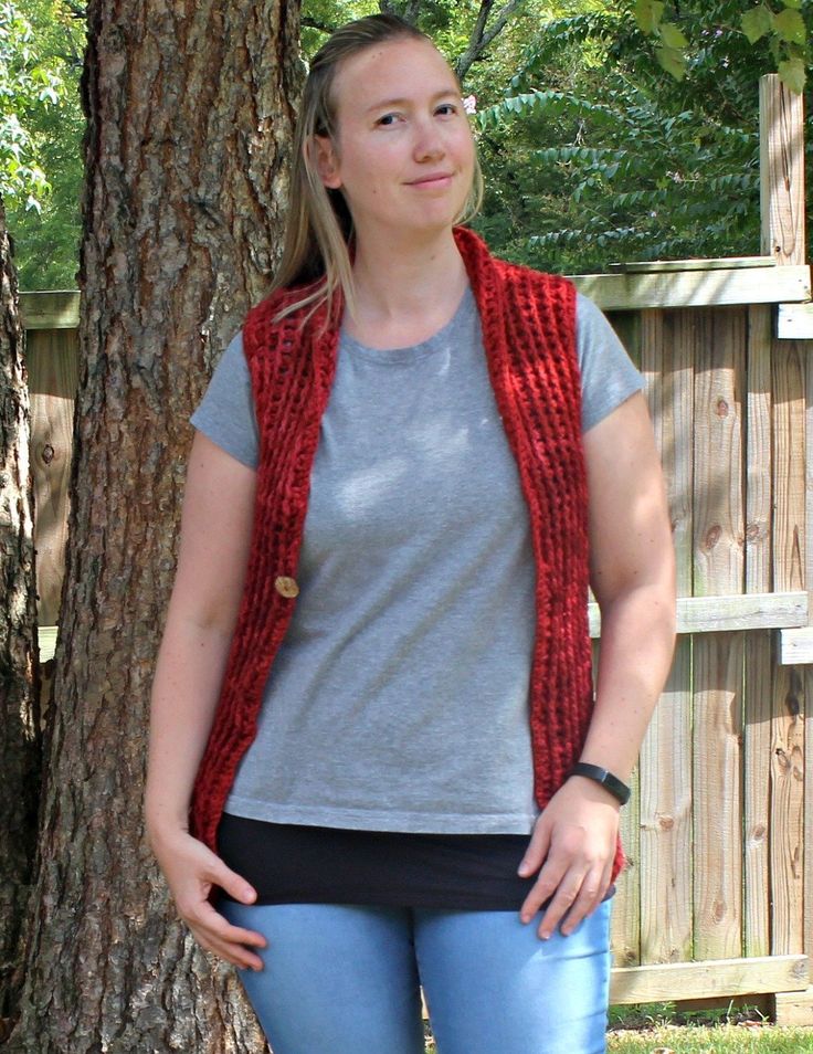 a woman standing in front of a tree wearing a red knitted vest and jeans