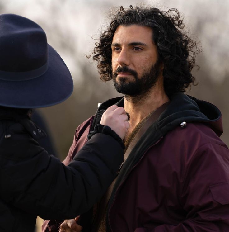 a man with long hair and a hat is talking to another man wearing a purple jacket