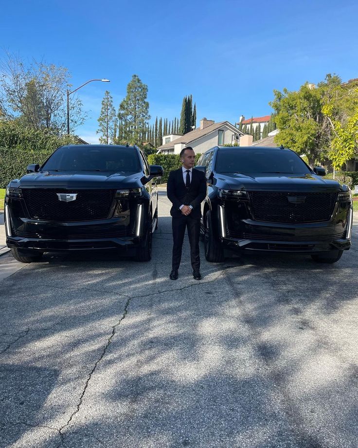 a man in a suit standing next to two black suvs on the street with trees behind them