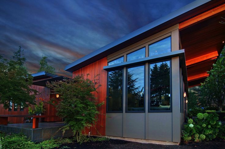 a house with large windows sitting next to trees and bushes at night, under a cloudy sky