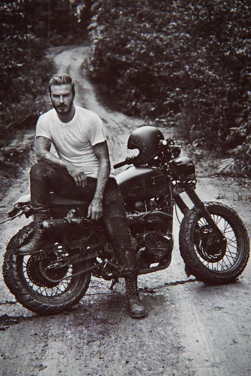 a man sitting on top of a motorcycle in the middle of a dirt road next to bushes