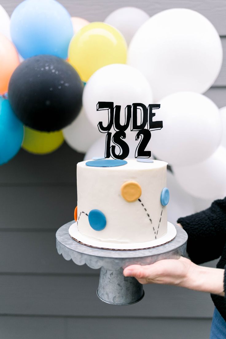 a person holding a cake with the number twelve on it and balloons in the background