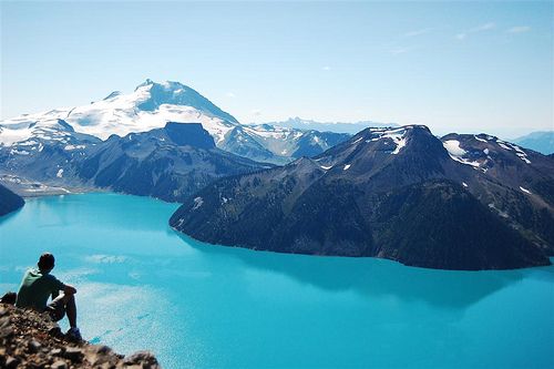 a man sitting on top of a mountain next to a large body of blue water