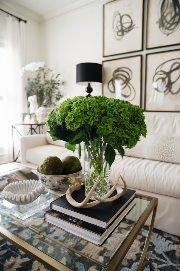 a glass table topped with green flowers and books next to a white couch in a living room