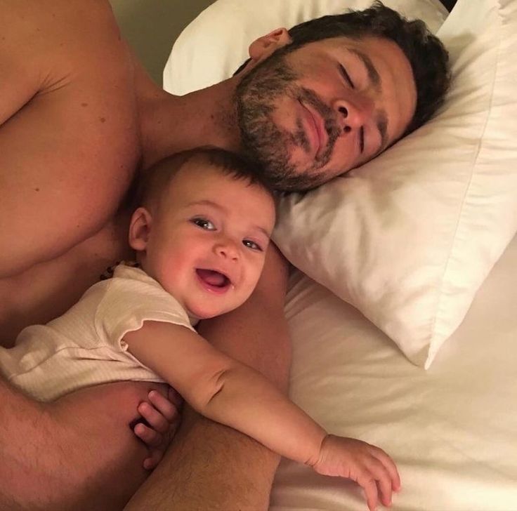 a man laying in bed next to a baby with his arm around the head of another man