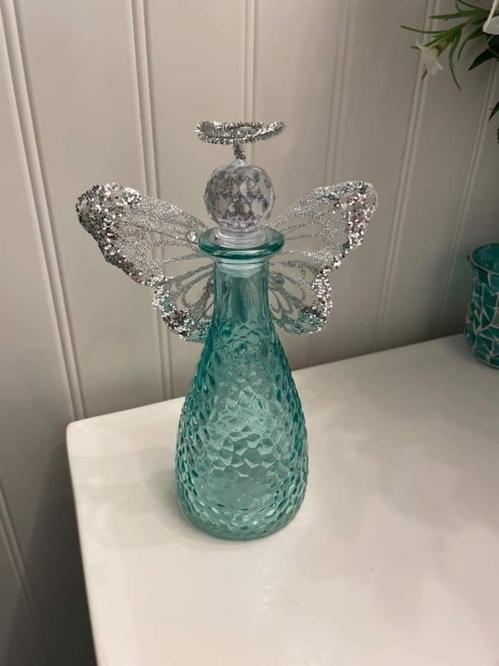 a glass bottle with an angel decoration on the top sitting on a white table next to a potted plant