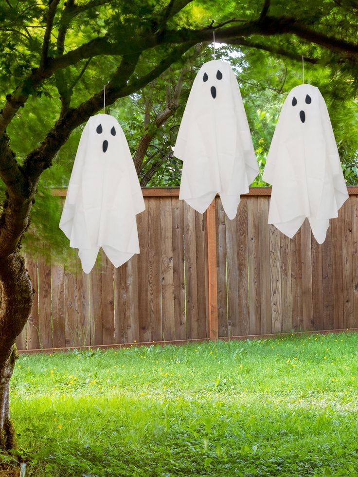 three ghost decorations hanging from a tree in front of a fence with grass and trees