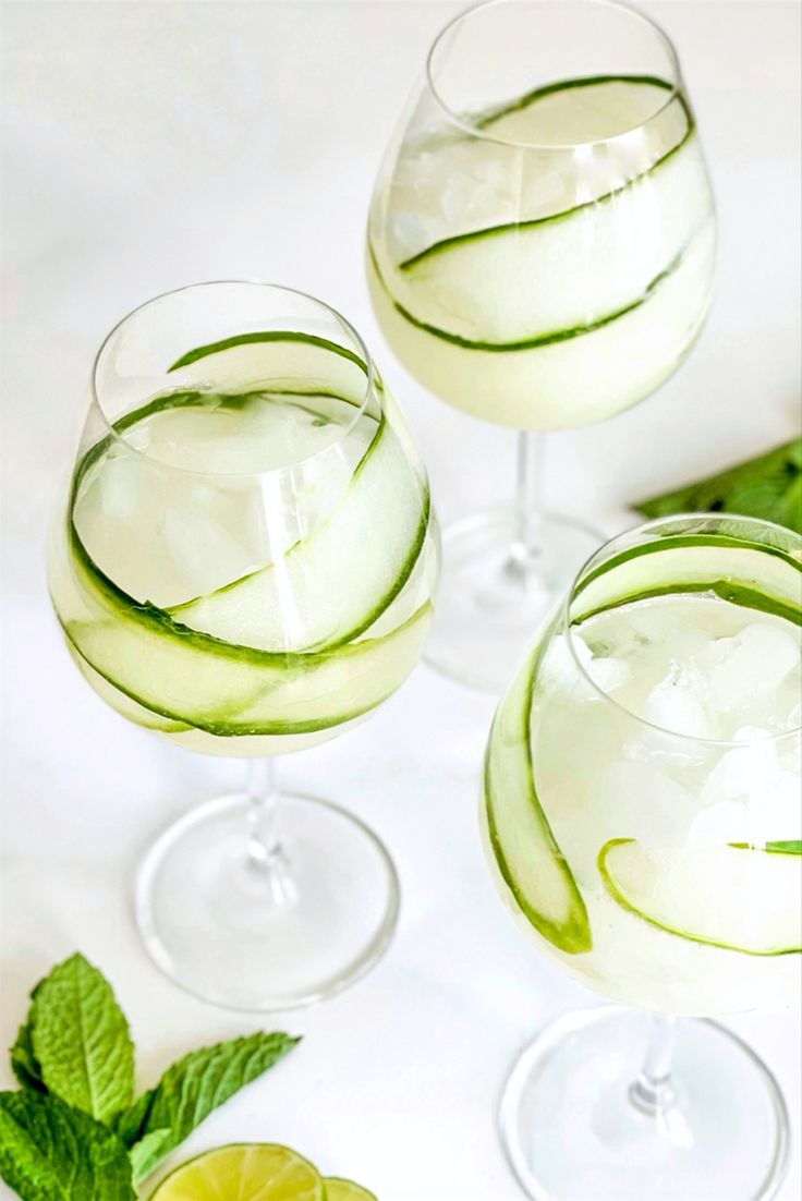 two glasses filled with cucumber and ice on top of a white tablecloth