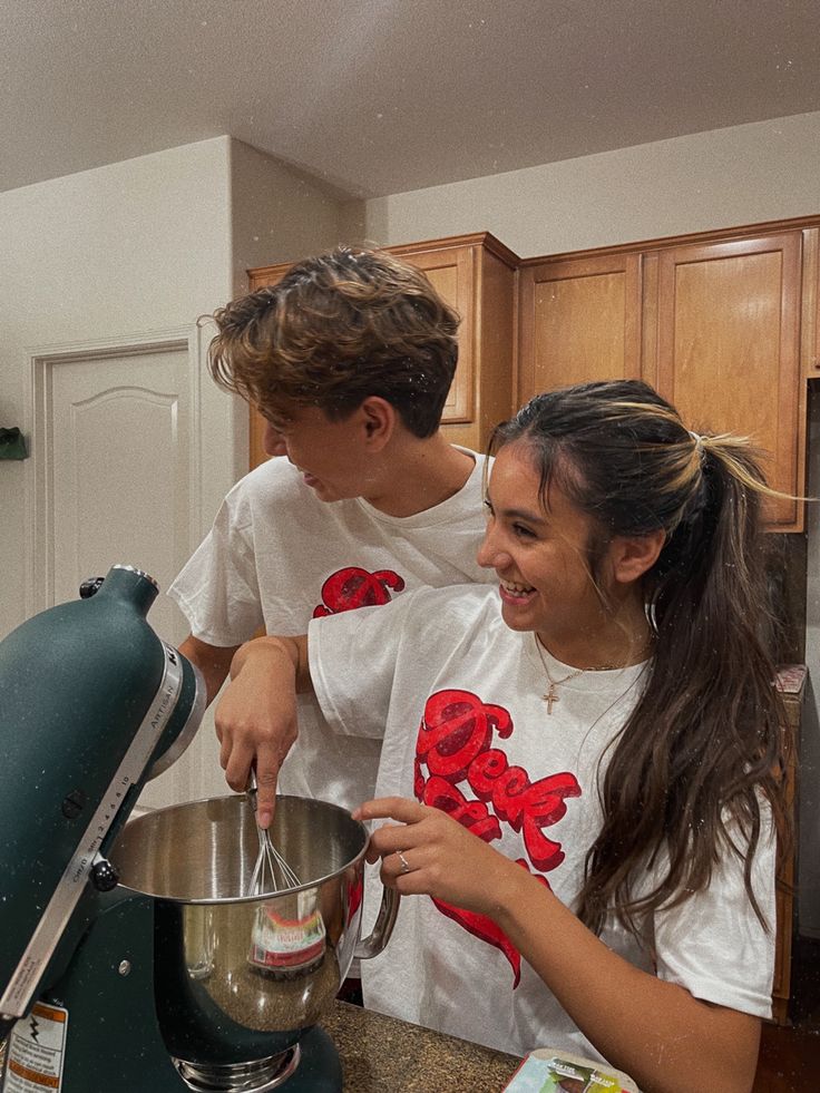 two young people are making something in a bowl on the kitchen counter, one is holding a mixer and the other is looking at it