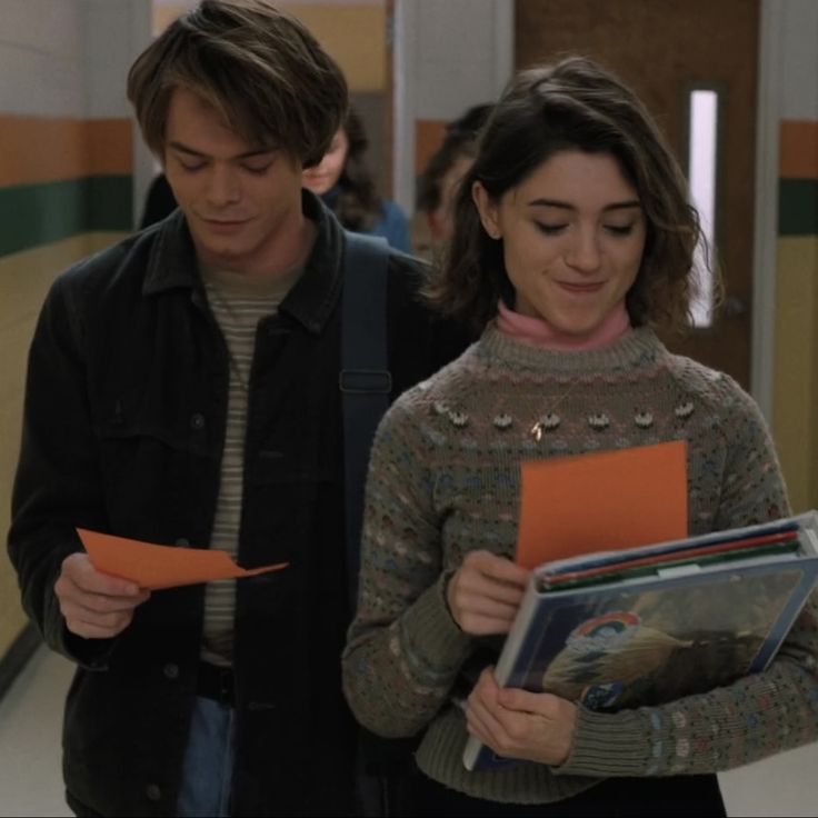 a man and woman walking down a hallway holding orange folders in their hands while looking at each other's papers