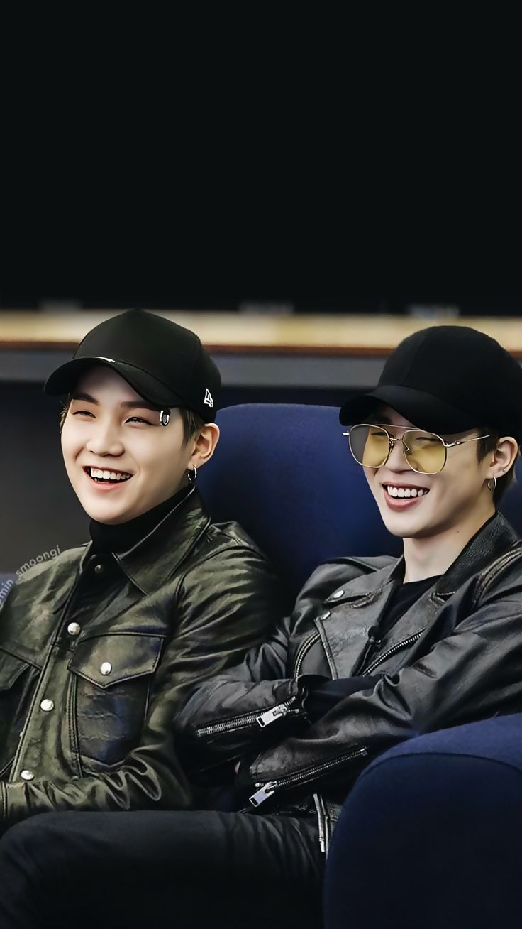 two young men sitting next to each other wearing black hats and leather jackets, one smiling at the camera