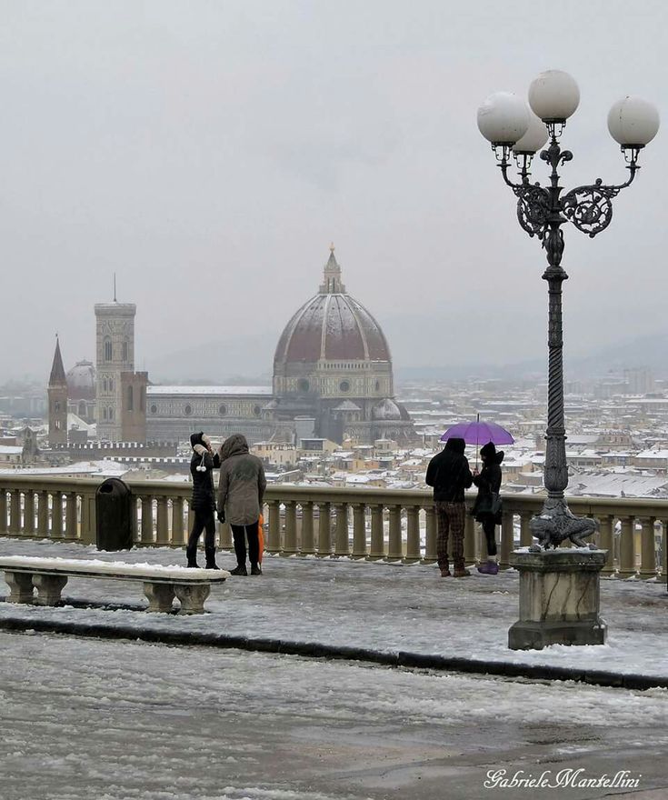 several people standing on a bridge with umbrellas in the snow and buildings in the background